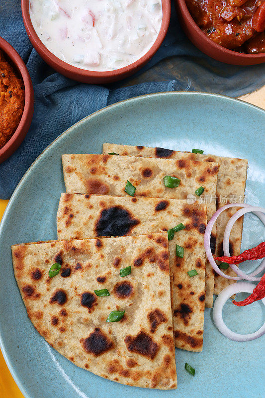 绿色的盘子，三角形的aloo paratha切片上撒有辣椒，红洋葱圈和切碎的韭菜，美味的面饼里塞满了捣碎的garam masala土豆，酸辣酱，泡菜蘸酱，raita(酸奶)，黄色的背景，俯瞰的风景